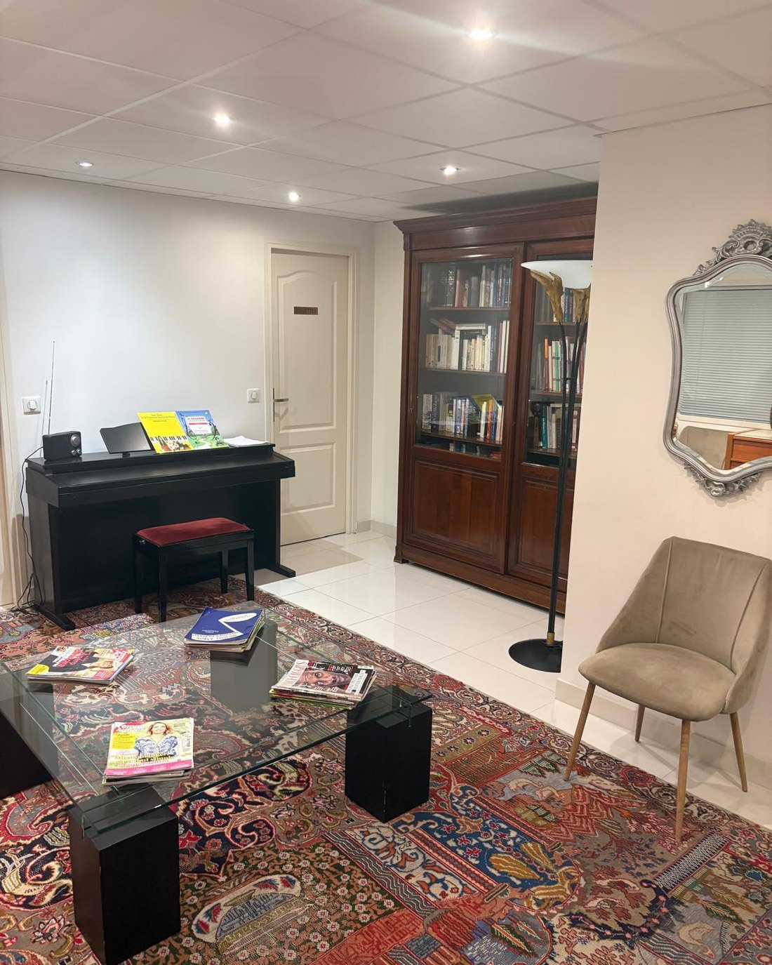 La salle d'attente du cabinet : un piano, une bibliothèque ancienne, un tapis et un fauteuil.